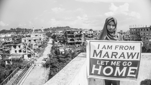 マラウィ旧戦闘地域の前で横断幕を掲げて写真で呼び掛けを行う同地域出身の若者=レット・ミー・ゴー・ホーム提供